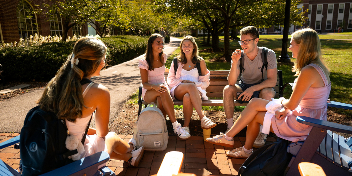 A group of students sits outside on benches in the sunshine. They are smiling.