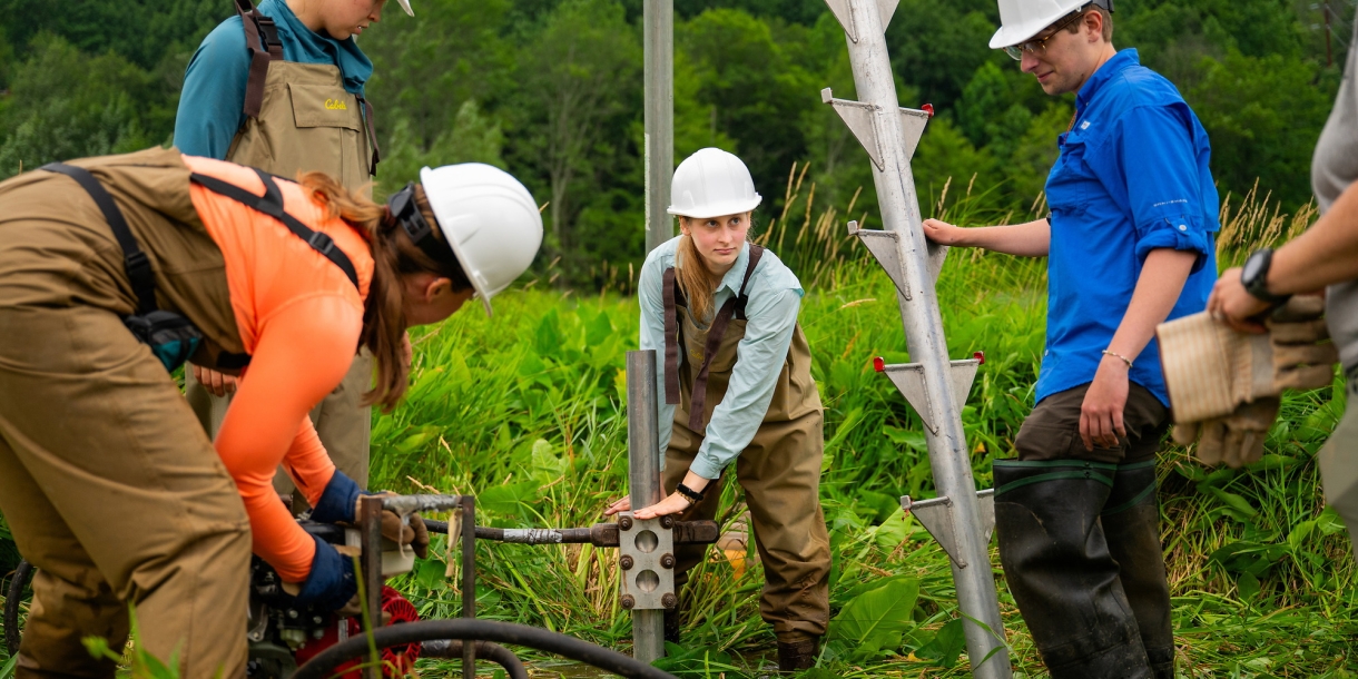 A group of Bucknell students wearing hardhats stand around a large machine in a field of green