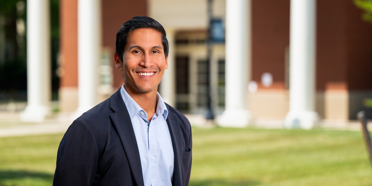 Professor Karlo Malaga stands on campus and smiles, a building with columns is in the background