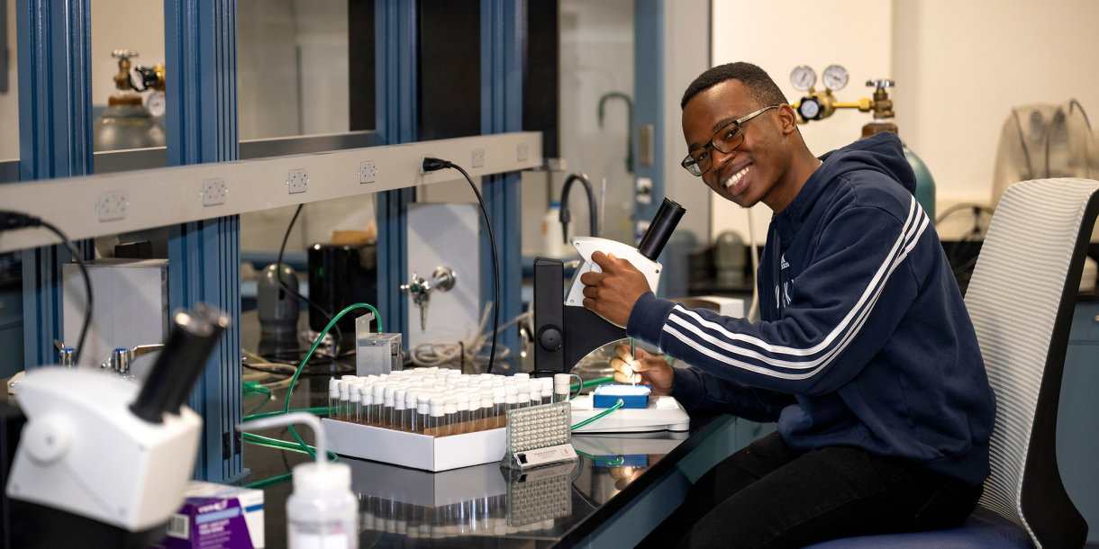 Monwabisi Qeki sits in a chair in a lab setting and smiles as he adjusts a microcope.