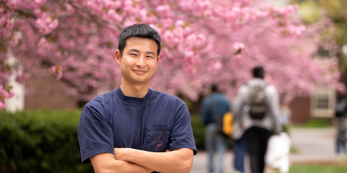 Tobey Kim wears a blue T-shirt and smiles on campus with cherry blossom trees behind him.