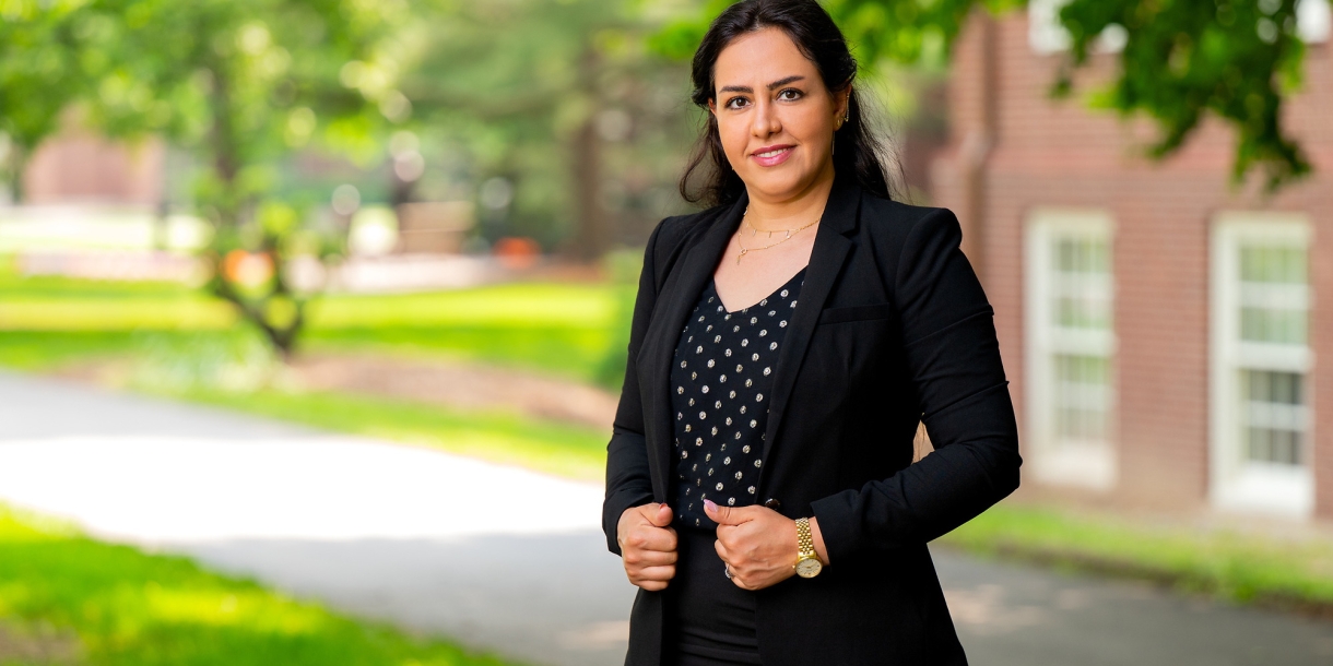 Vaji Farhadi wears a black suit and smiles while standing outside on campus with green trees and sunshine behind her.