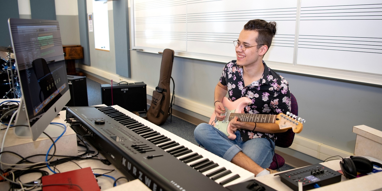 Ian Herdt plays a guitar in front of a musical keyboard and computer