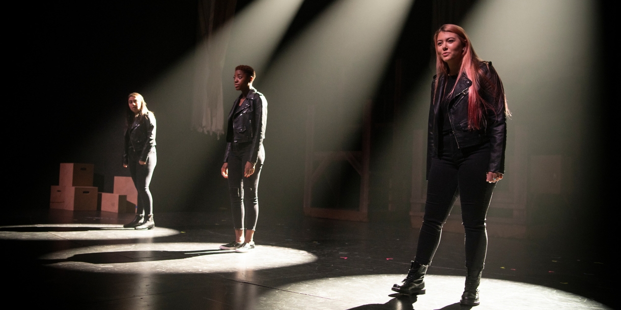 Three actresses stand on stage under spotlights.