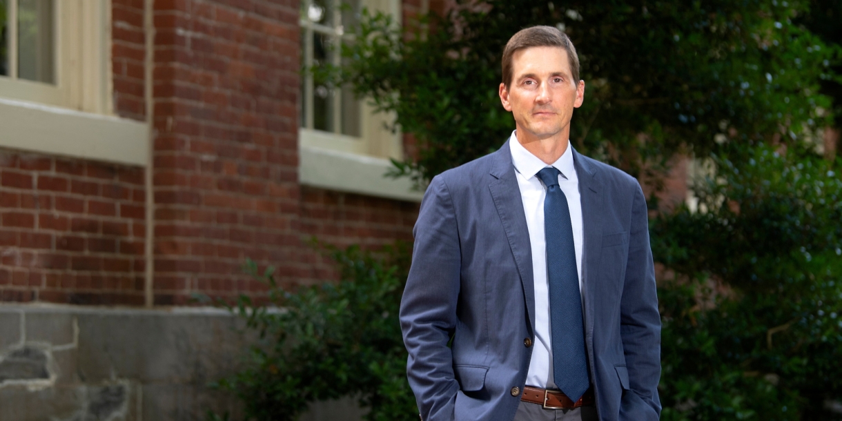 Professor Matt Bailey, in a blue jacket and tie, stands beside a brick building on Bucknell&#039;s campus