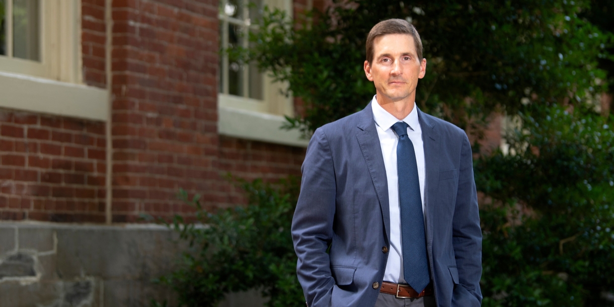 Professor Matt Bailey, in a blue jacket and tie, stands beside a brick building on Bucknell's campus