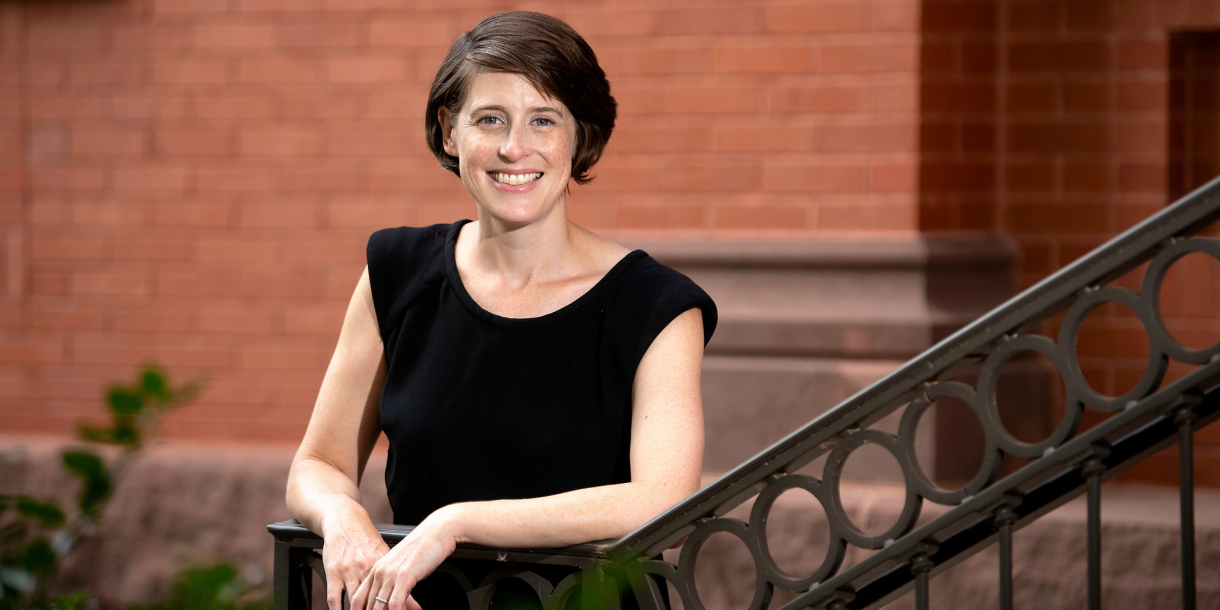 Reggie Gazes wears a black sleeveless shirt and stands against the railing of a staircase outdoors on campus with a red brick building behind her.