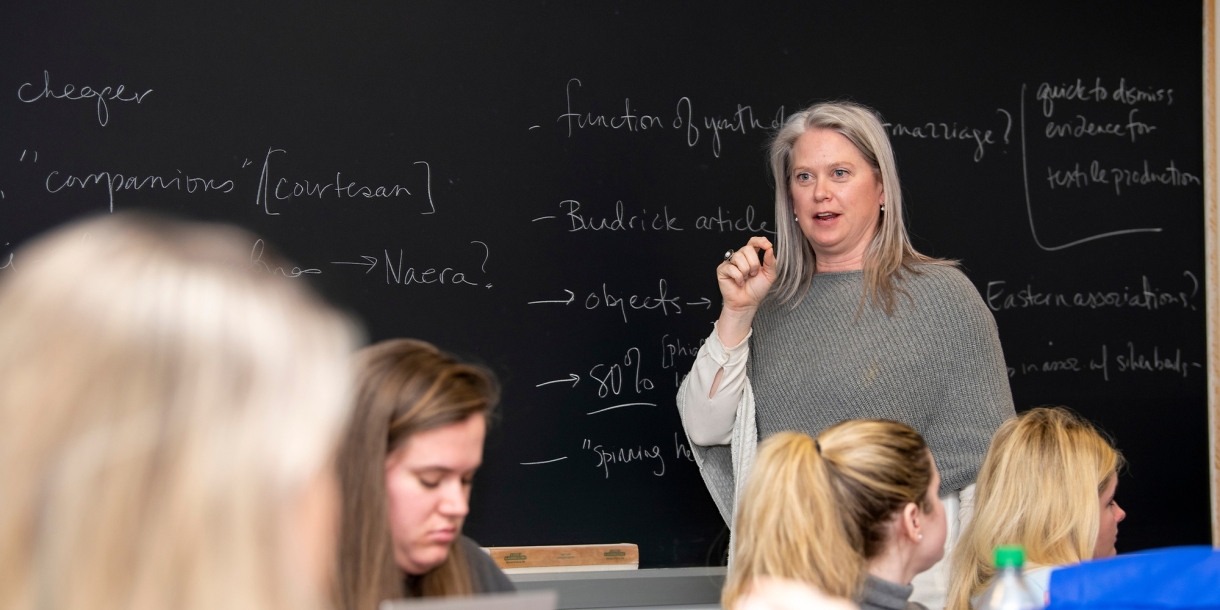 Professor Stephanie Larson stands in front of a chalk board while teaching a Women in Antiquities class.