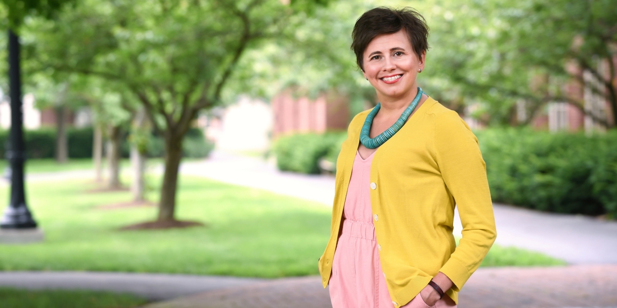 Erica Delsandro wears a yellow sweater over a pink dress and smiles on campus with sunlight shining through trees behind her.