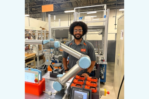 Kellen Haile stands in a factory-like setting wearing safety goggles and standing behind a large robotic arm.