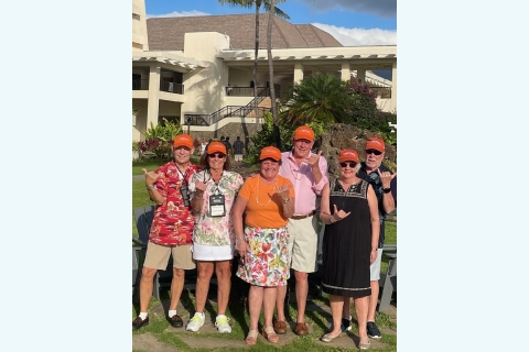 A group of alumni stand in front of a resort building orange hats and giving the "hang 10" hand sign.