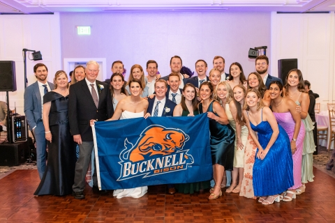 a large group of Bucknellians gather at a wedding, with the bride and groom in the center holding a Bucknell Bison flag.