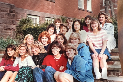 a group of students from the 1990s sit together on campus steps and smile.