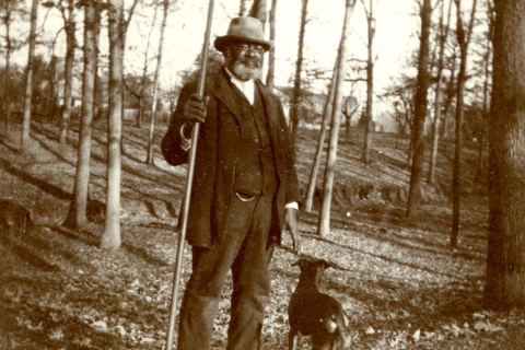 An archival photo shows Charles Bell, a tall Black man, standing with his dog among trees.