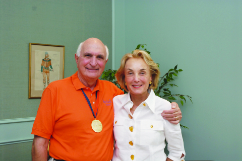 A man in an orange polo shirt poses with a woman in a white button-down blouse. He wears a gold medal around his neck. Both are simling.