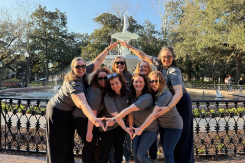 a group of women gather in front of a large fountain and smile with outstretched arms.