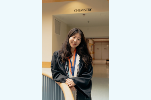Lucy Park wears a graduation gown with honor chords and stands inside a campus building with a "chemistry" sign behind her.