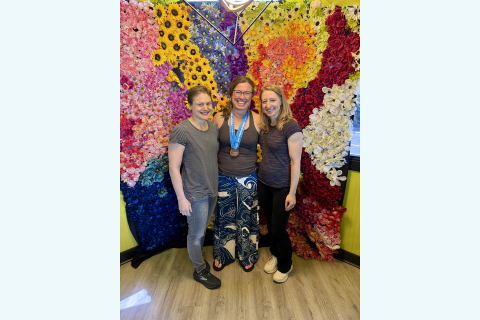 Three women stand and smile against a wall of colorful flowers; the woman in the center is wearing a medal around her neck.