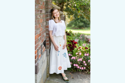 Ibby New wears a white dress while leaning against a brick wall