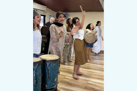 A group of barefoot performers sing, clap and play drums in a classroom. One performer raises a drumstick while others sing behind her.