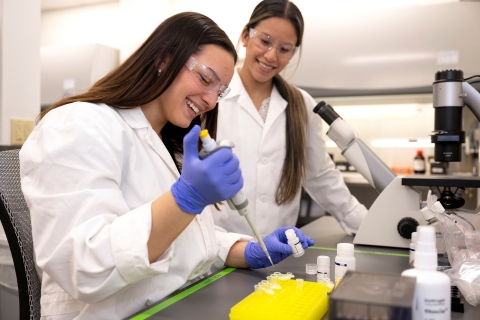 Elizabeth Malley '26 conducts lab work while wearing safety goggles and a white lab coat as Professor Olivia Boerman looks on and smiles.