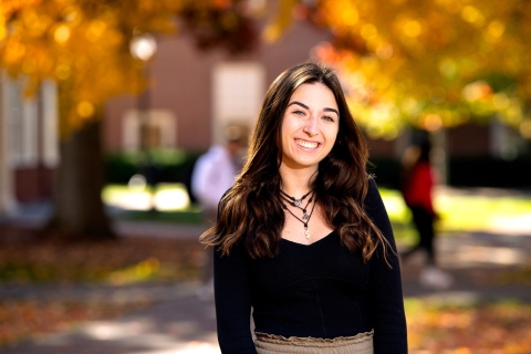 Elle Chrampanis &#039;24 smiles on campus, fall foliage is visible behind her.