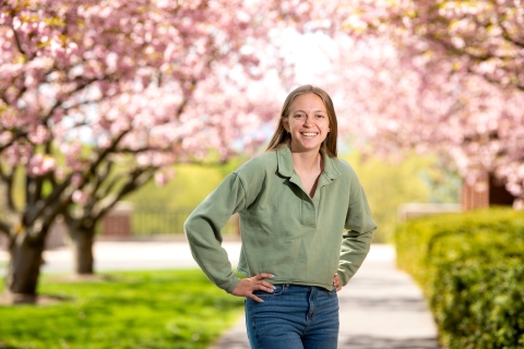 Ally Clarke &#039;24 smiles and stands on a campus pathway with both hands on her hips, pink cherry blossoms fill the backdrop