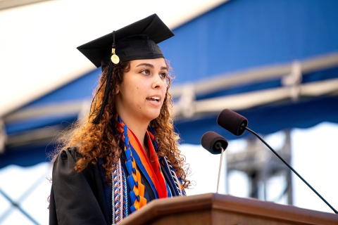 Kaia Rendo &#039;23 wears a cap and gown and honor chords as she speaks into a microphone at a podium at Commencement.