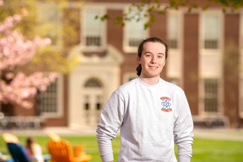Ethan McNamera wears a Bucknell chemistry sweatshirt and smiles on campus, with a red brick building and cherry blossoms behind him