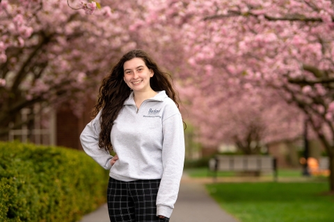 Patricia Poley &#039;23 stands with one hand on her hip on a campus pathway with pink cherry blossoms behind her.