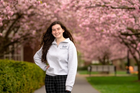 Patricia Poley '23 stands with one hand on her hip on a campus pathway with pink cherry blossoms behind her.