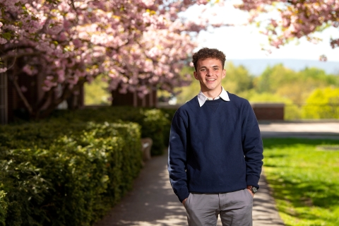 Sam Douds wears a blue sweater and gray pants while smiling and standing on a campus pathway with cherry blossoms behind him