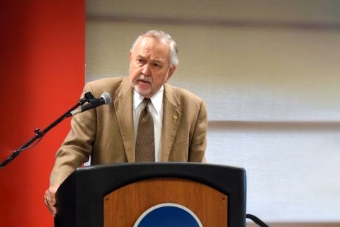 Merrett Stierheim wears a suit and stands behind a podium and speaks into a microphone.