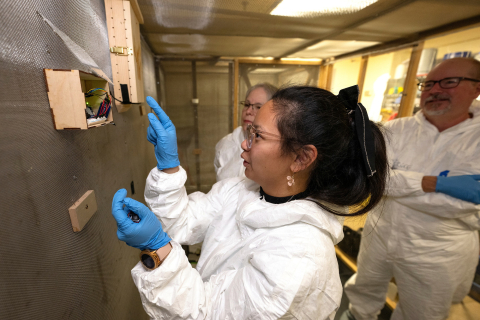 Angel Eunisse Yanga wears blue gloves and a white safety suit as she points at a device on the wall as professors Ken Field and DeeAnn Reeder look on from behind her.