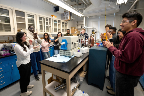 My Ly holds an award in a lab setting and is surrounded by a group of about 10 student engineers and a professor who are smiling and applauding.