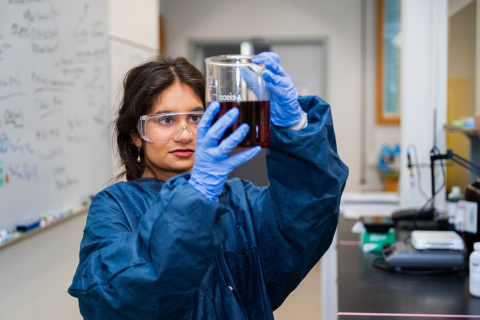 Priyanka Poplai '29 wears a blue lab coat, blue gloves, and safety goggles while she holds up a liquid in a clear container in a lab space.