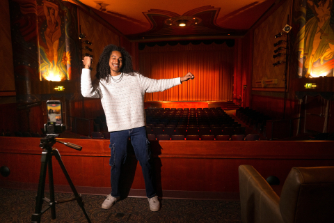 Caleb Warren stands in front of his phone and poses with one arm up; his phone is on a tripod and is arranged so it is filming him.