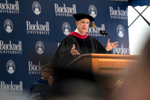 Ken Freeman wears black graduation robes while speaking at a podium.