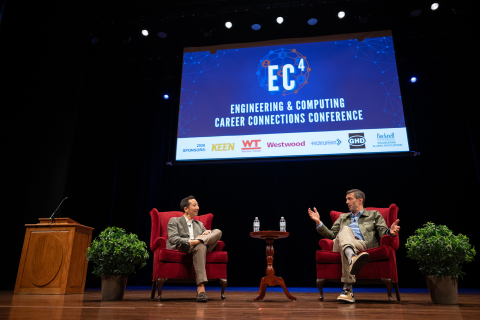 Jordan Rice gestures while speaking with Professor Charles Kim. Both are seated in red armchairs onstage beneath a screen displaying the EC⁴ conference name.