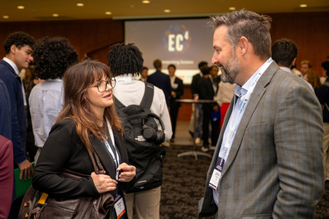 A student wearing glasses talks with a smiling guest in a gray blazer as other attendees network behind them beneath an EC⁴ projection.