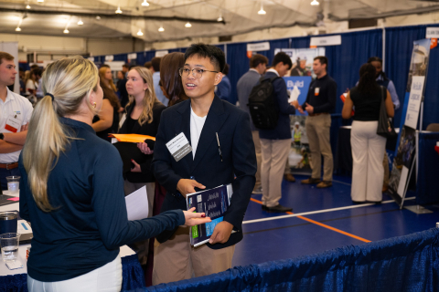 A student holding informational materials speaks with an employer representative while other attendees talk at booths throughout a busy career fair.