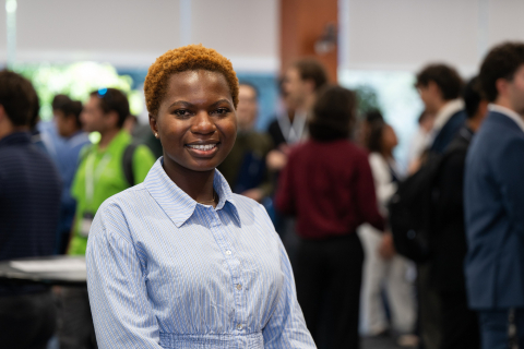 Simbi Maphosa smiles at the camera in a blue-and-white striped shirt, with conference attendees gathered behind her.
