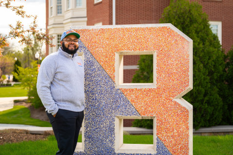 Jawaad "KJ" Benson stands next to a large letter B that is part blue and part orange.