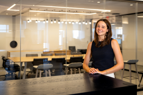 Kelly Byrne stands at the end of a dark high-top table; an office scene is in the background