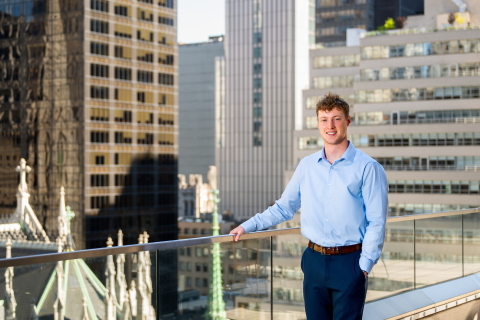 Max Wilson stands outside on a rooftop balcony with his hand resting on the railing; city buildings are behind him.
