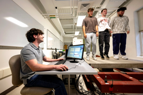 Michael Evanick sits at a desk with a computer as classmates stand on a platform to his left.