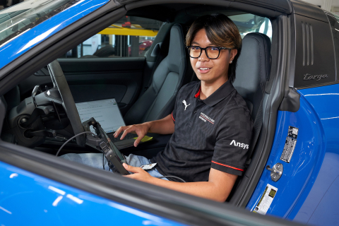 Zachary Ebilane sits in the driver's seat of a bright blue car with a laptop open on the passenger side seat.