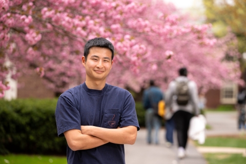 Tobey Kim wears a blue T-shirt and smiles on campus with cherry blossom trees behind him.