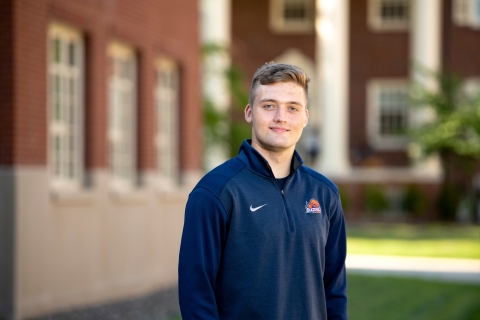 Duncan Hill wears a Bucknell Bison blue long-sleeved shirt and stands outside on campus with red brick buildings in the background.