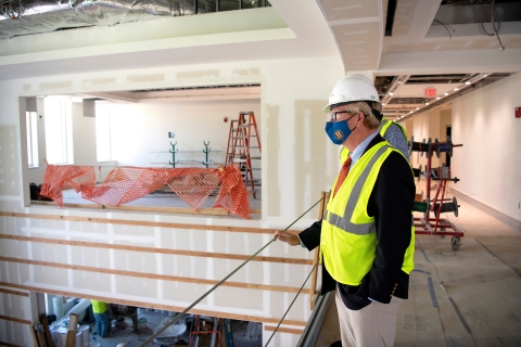 Wearing a hard hat, mask and high-visibility vest, Steve Holmes looks over a balcony at the under-construction Holmes Hall.